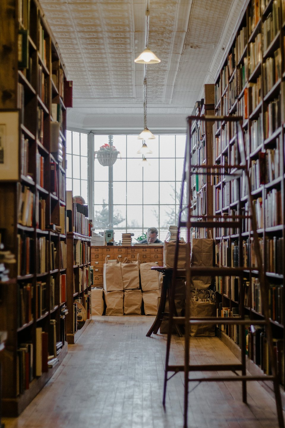 Bookstore aisle with wooden shelves leading to a bay window where someone is reading.